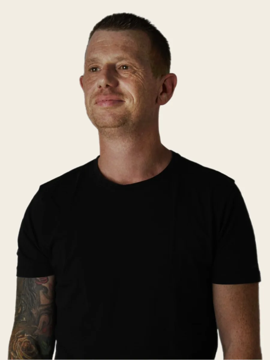 Portret van Brian van Straalen, in zwart T-shirt met een tatoeage op zijn onderarm, tegen een effen achtergrond.