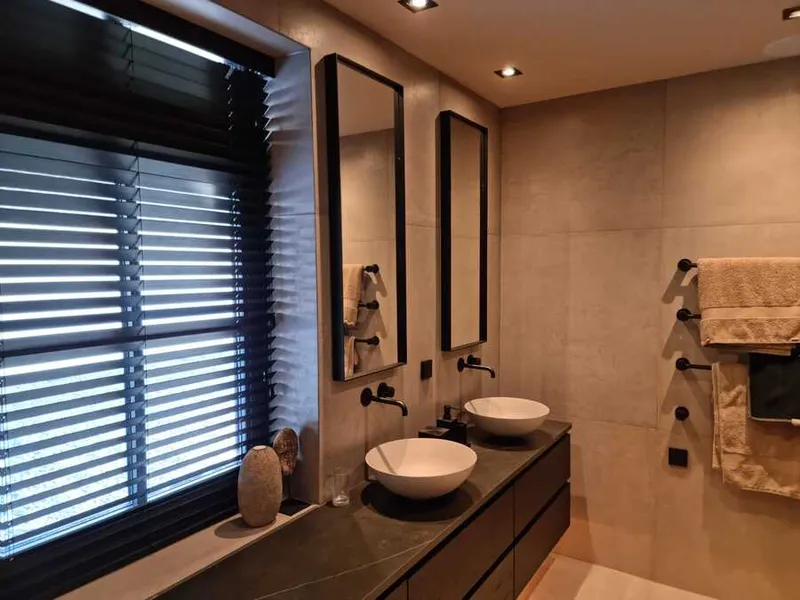 Gerenoveerde badkamer met een donker wastafelmeubel en twee witte waskommen, zwarte kranen en spiegels, en wanden in een warme zandkleur.