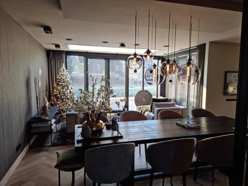 Eethoek met een donkere wand, een houten tafel met stoelen en hanglampen van glas voor het raam.