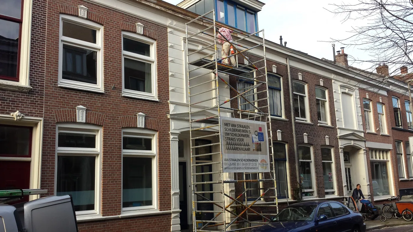 Straat met bakstenen herenhuizen; tegen een witte gevel staat een rolsteiger met een schilder erop en een bouwbord van Van Straalen Schilderwerken.