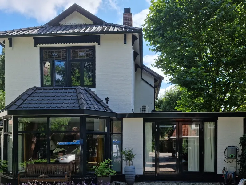 Wit geschilderd woonhuis in Hilversum met zwarte kozijnen, een serre met een pannendak en een aanbouw met openslaande deuren, voor een grindpad en lavendel.
