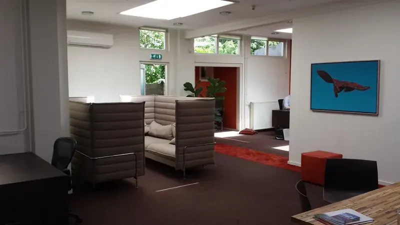 Kantoorruimte met wit gelatexte wanden en plafond, grijze loungebanken en een rood tapijt.