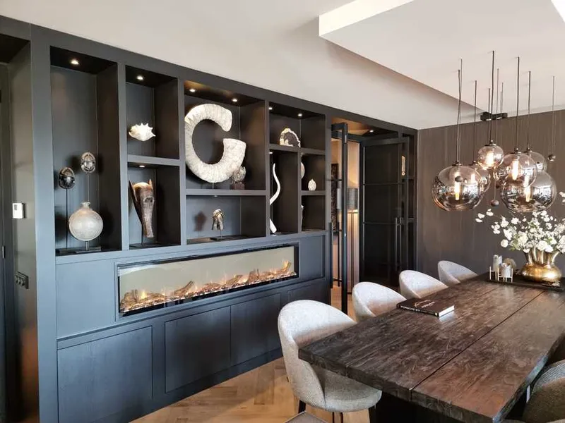 Eetkamer met een donkere kastenwand van vloer tot plafond: open vakken met spotjes erboven en een brede ingebouwde haard eronder.