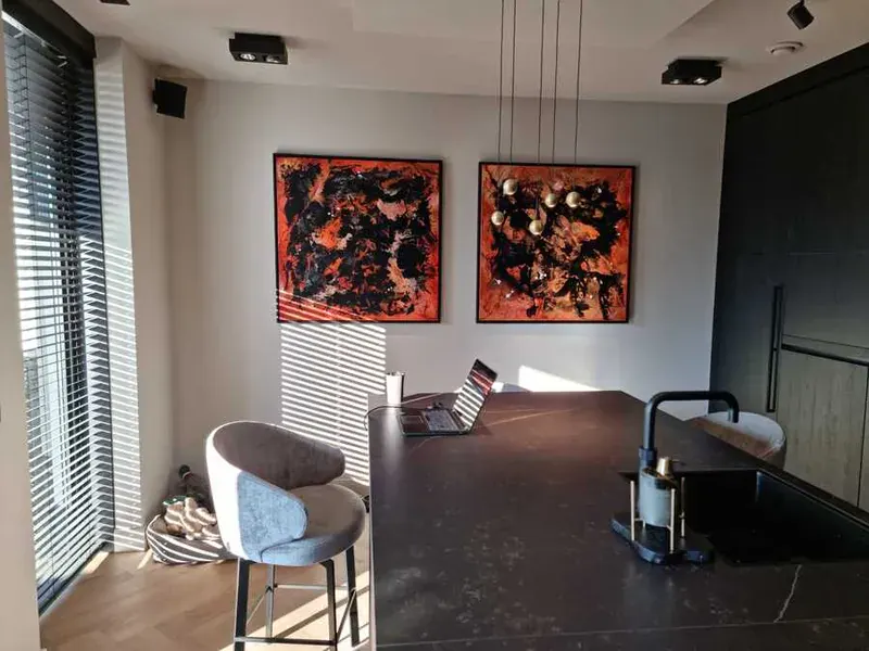 Keuken met een donker kookeiland en twee oranje-zwarte schilderijen aan een lichte wand.