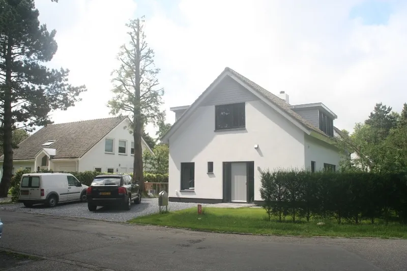 Vrijstaand wit geschilderd woonhuis met donkere kozijnen en een puntdak, aan een straat met hoge bomen.