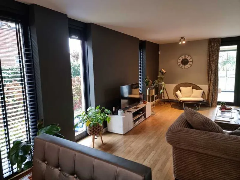 Woonkamer met bruingrijze wanden, hoge ramen met lamellen, een donkere houten vloer en een lichte bank.