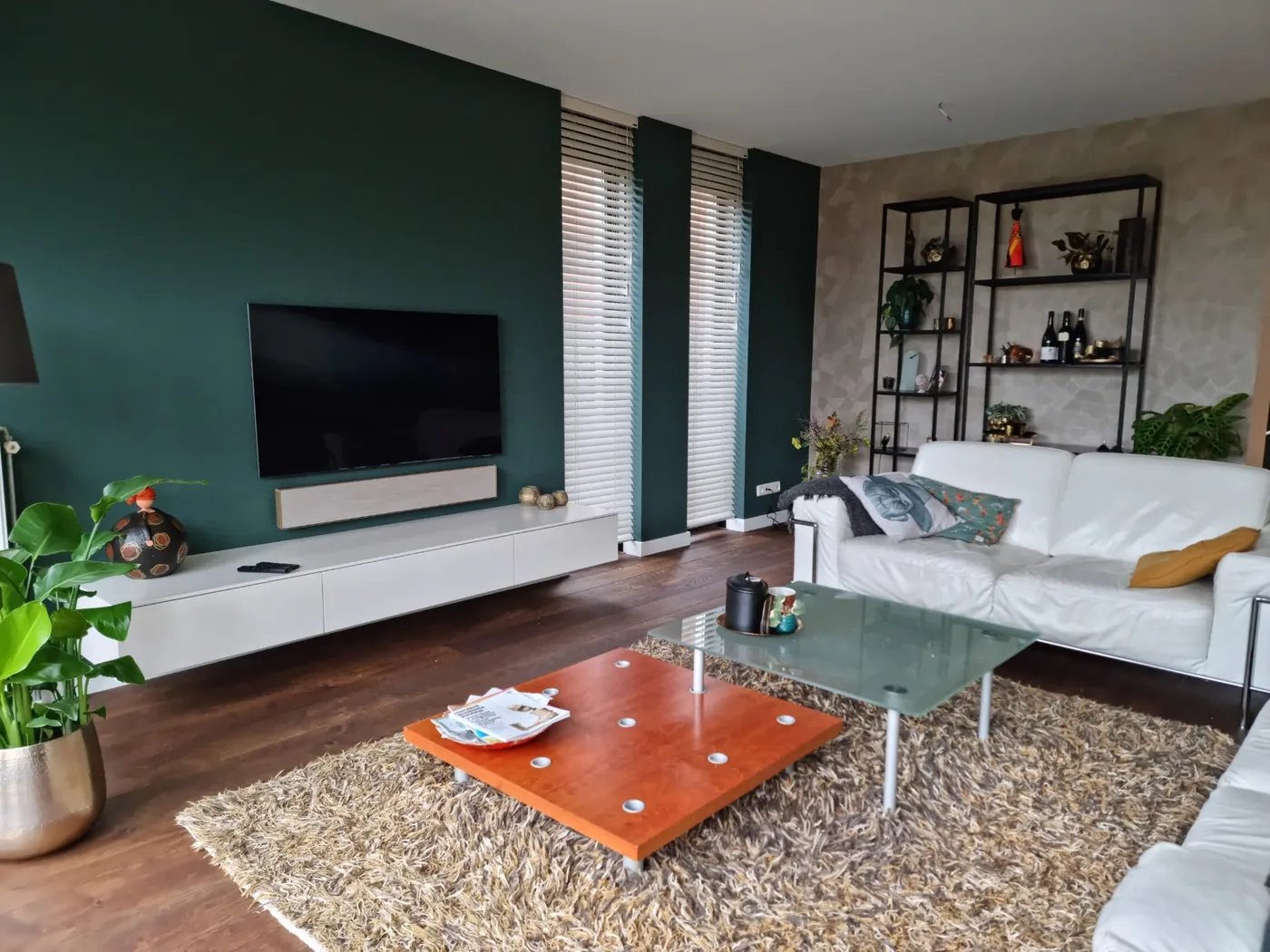Lichte woonkamer met een donkergroene wand achter de televisie en twee donkergroene penanten tussen de ramen; op de vloer een witte leren bank en een oranje salontafel.