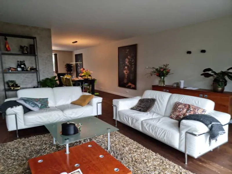 Lichte woonkamer met witte wanden en plafond, twee witte leren banken en een oranje salontafel.