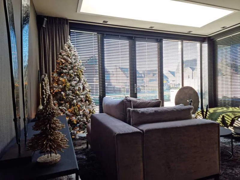 Zithoek met een grijze bank, houten lamellen voor de ramen en een kerstboom met lichtjes in de hoek.